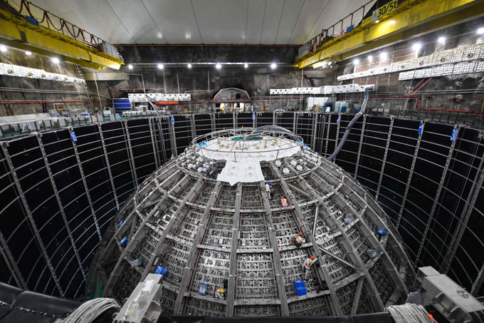 An experimental hall houses a large spherical apparatus with workers climbing on it.